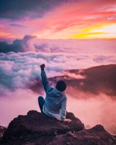 person in jean jacket sitting on the top of a mountain with fist up in success. Sunset in the background